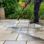 Load image into Gallery viewer, A person wearing dark work pants and red gloves uses a pressure washer to clean a stone paver patio in a garden setting.